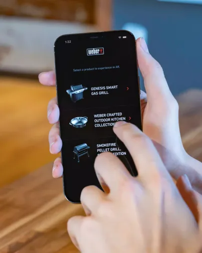 Right hand holding phone with Weber AR homepage open and left hand raised to touch in to application