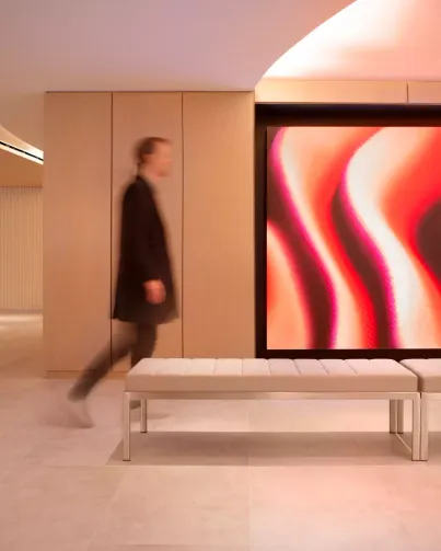 A modern interior features a person walking past a vibrant, abstract digital art display with red and orange waves. The space has light wood panels and a sleek white bench, creating a clean, contemporary look.