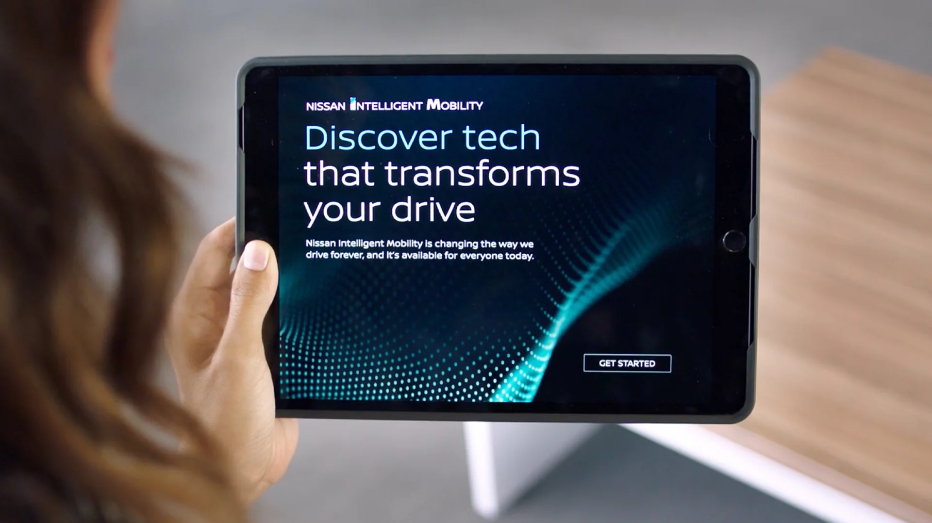 A person holding a tablet with the text "Nissan Intelligent Mobility. Discover tech that transforms your drive. Nissan Intelligent Mobility is changing the way we drive forever, and it's available for everyone today."