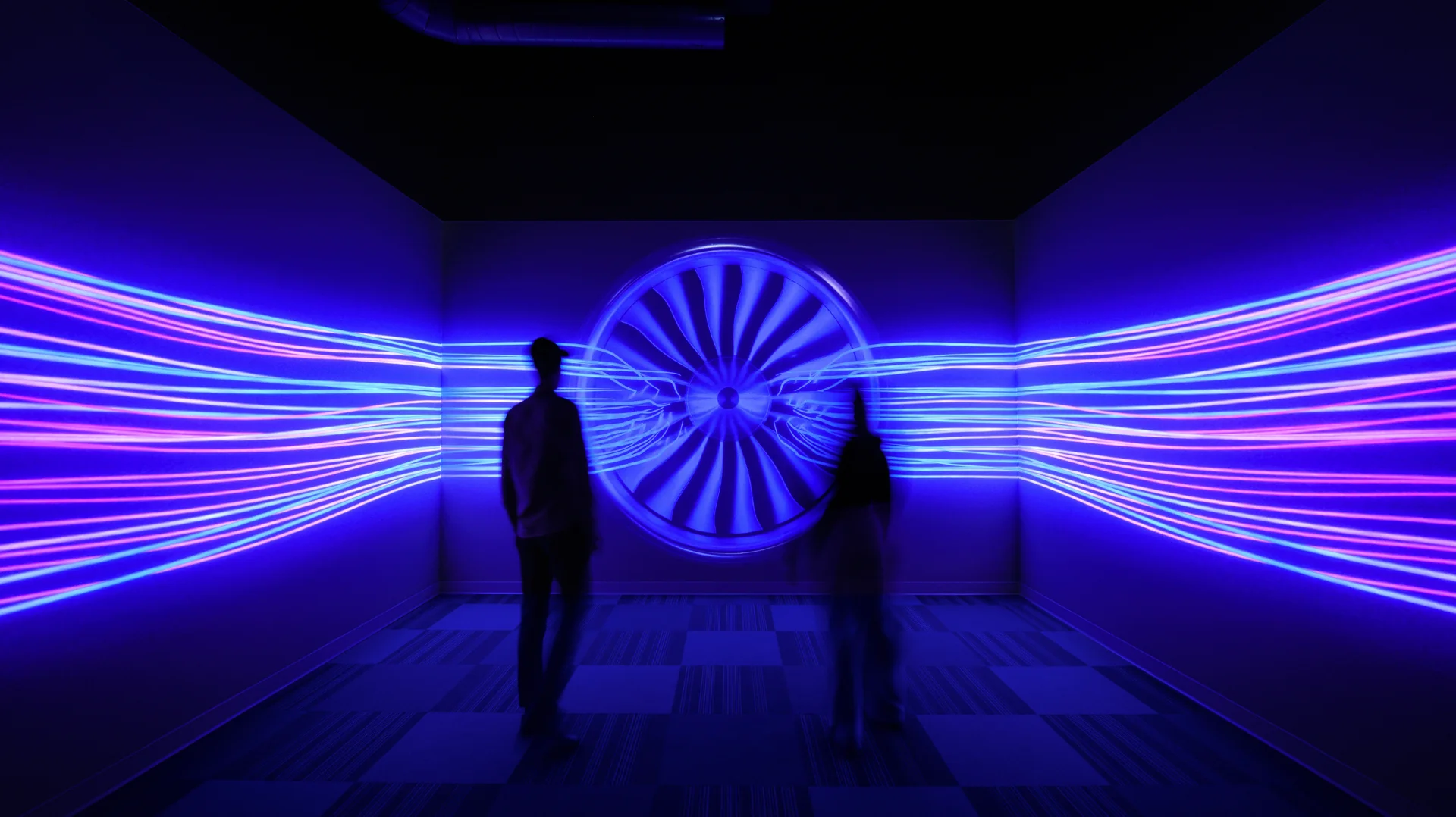 Two people standing in the center of a small room with a glowing blue image of an airplane engine covering all three walls