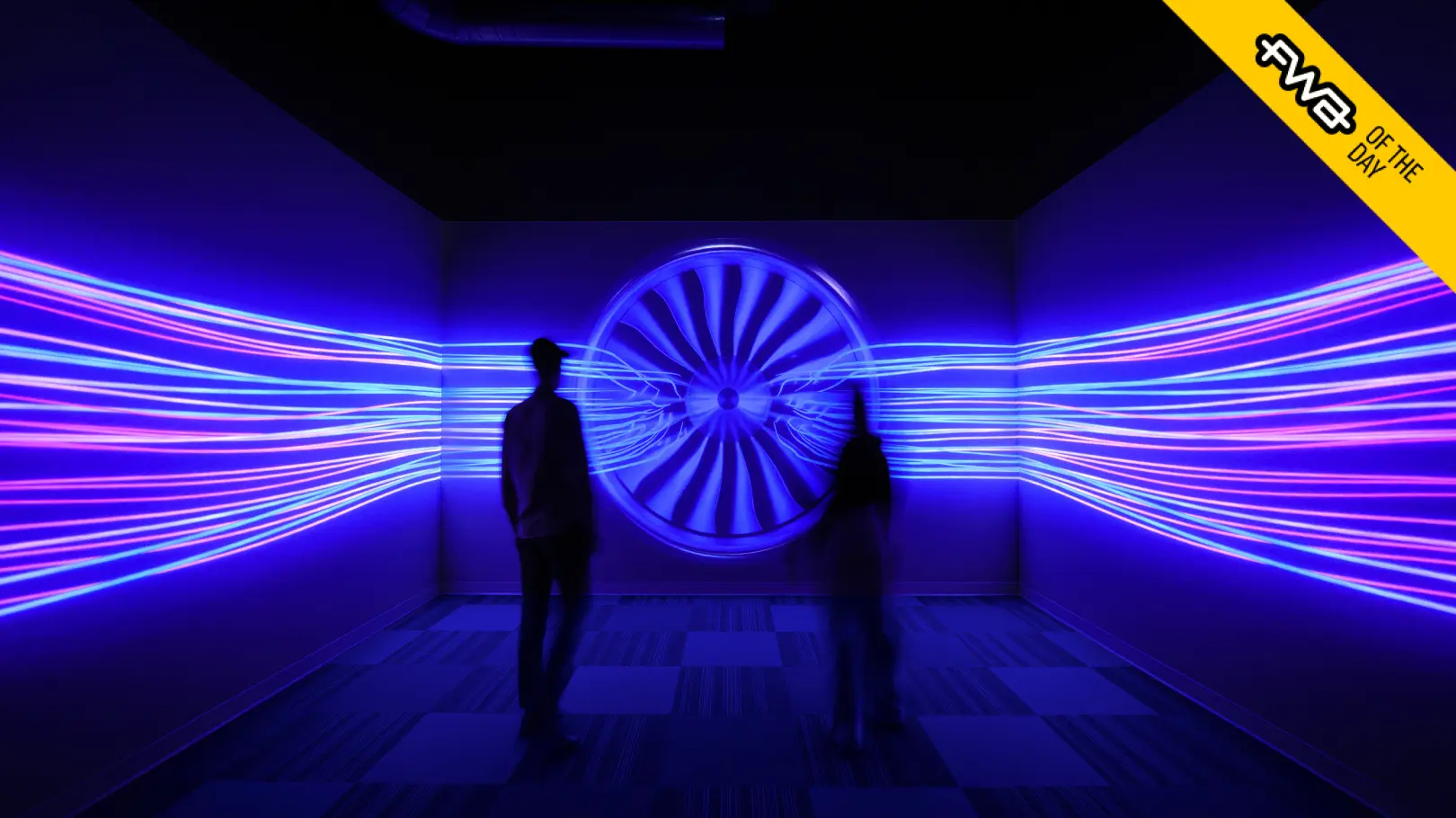 Two people standing in the center of a small room with a glowing blue image of an airplane engine covering all three walls, overlayed with "FWA of the Day" banner